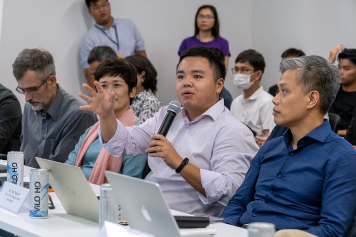 The judges panel gives feedback. A man with a purple shirt holds the mic and has a hand raised while describing his thought. 