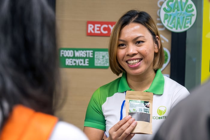 A woman with dark hair and blonde highlights holds a sealed bag of compost. She wears a green and Eco Vietnam shirt and stands in front of a display with signs that say "Food Waste Recycle" and "Save the Planet". 