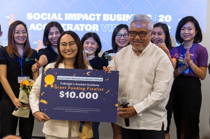 A woman with glasses and a man with glasses both wear white and are holding an award for the Grant Funding Finalist. A line of clapping people stand behind them, and they all stand in front of a projected slideshow for the Social Impact Business Accelerator. 